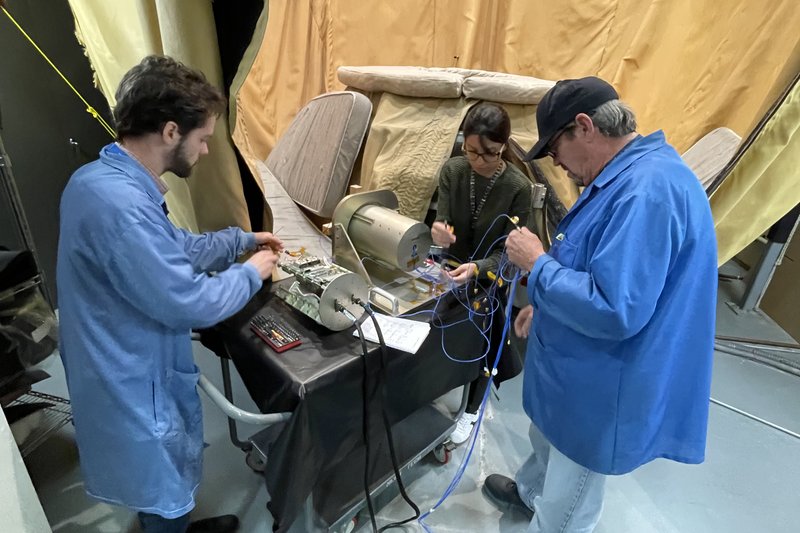 Three engineers, two in blue lab coats and one in casual clothing, work on a piece of scientific equipment on a cart. The equipment is surrounded by various wires and tools, and the team members appear focused on their work. The background features a large, draped curtain.