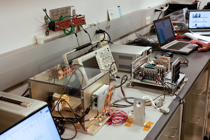 The image shows a laboratory workspace with various electronic testing and measurement equipment on a countertop. A nearby laptop displays data, and colorful wires connect the devices.